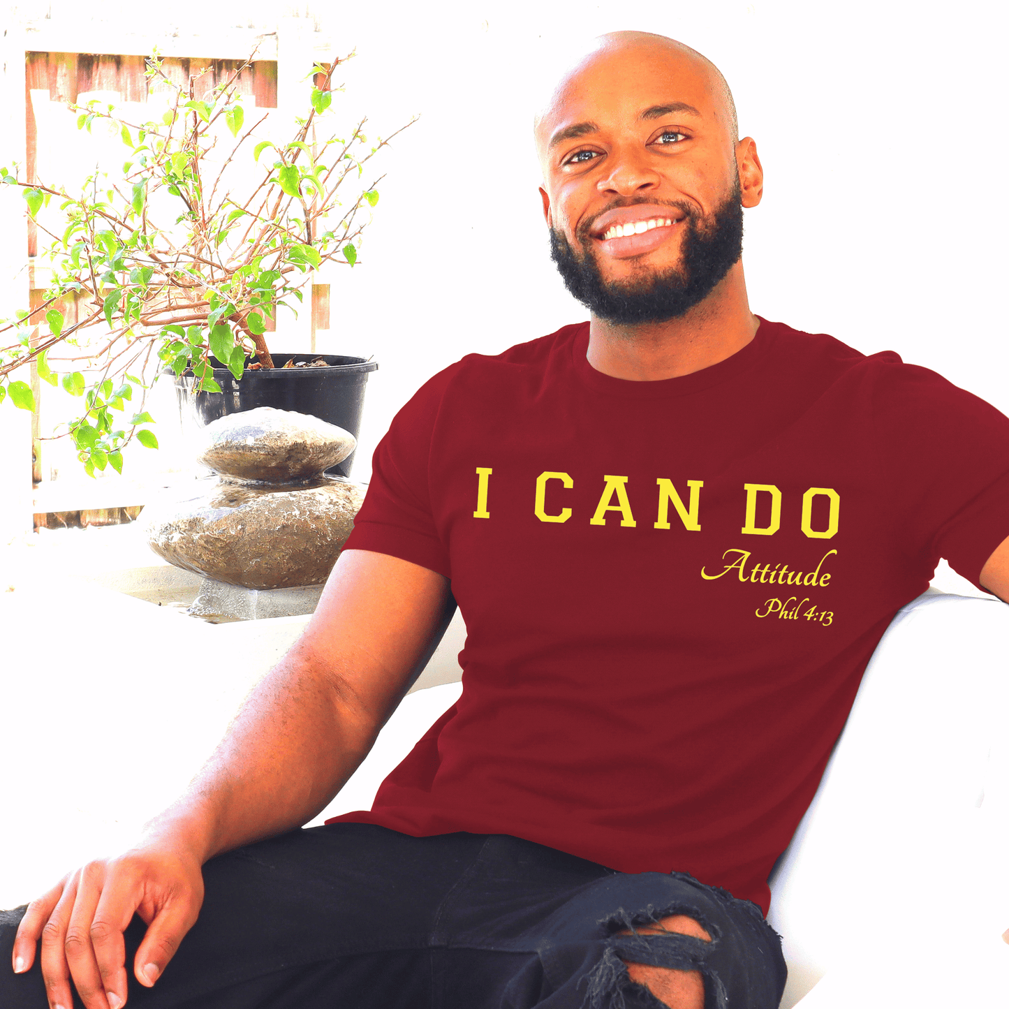 Man wearing a maroon t-shirt with 'I CAN DO Attitude Phil 4:13' text,by marto and tshirts, sitting in a room with plants.